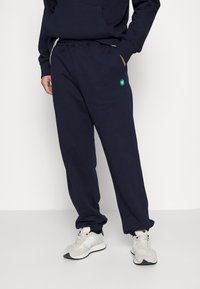 Pantalones de chándal azul marino con cintura elástica, bolsillos laterales y un detalle de logo verde. Fabricados con una tela suave y con un ajuste relajado.