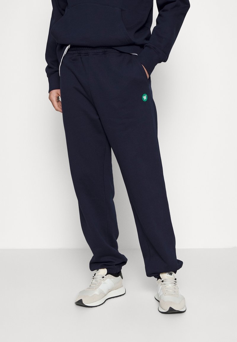 Pantalones de chándal azul marino con cintura elástica, bolsillos laterales y un detalle de logo verde. Fabricados con una tela suave y con un ajuste relajado.