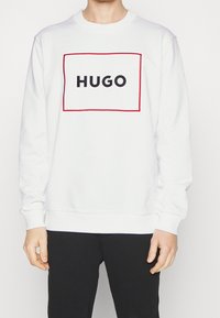 Valkoinen sweatshirt, jossa on pyöreä kaula-aukko, punainen suorakulmainen reunus ja sana "HUGO" rohkeilla mustilla kirjaimilla rinnan kohdalla.