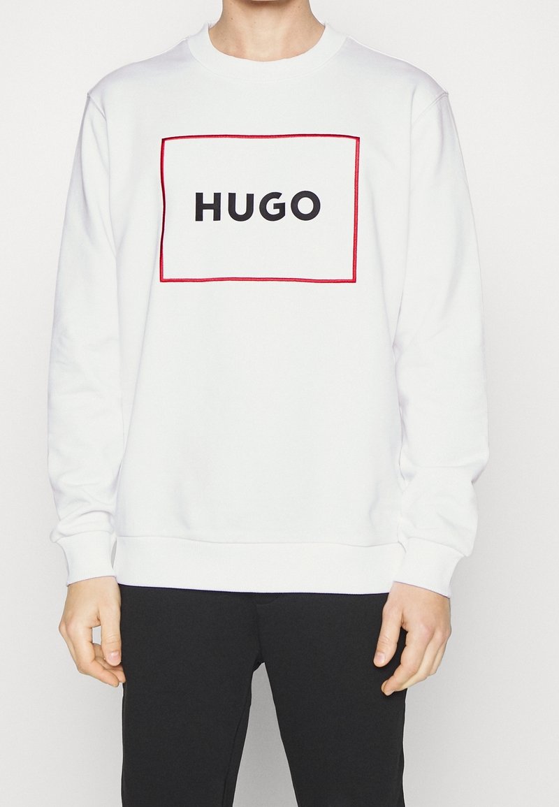 Valkoinen sweatshirt, jossa on pyöreä kaula-aukko, punainen suorakulmainen reunus ja sana "HUGO" rohkeilla mustilla kirjaimilla rinnan kohdalla.