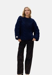 Gruby sweter w kolorze granatowym w splot kabelkowy o szerokiej sylwetce, z teksturowanymi rękawami i małą metką z logo po boku, zestawiony z ciemnymi, luźnymi spodniami.