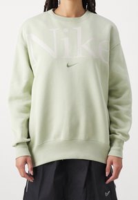 Ljusgrön Nike-hoodie med oversize-passform, stor rosa logotyp på framsidan, ribbade ärmslut och nederkant, mjuk textur, bärs med svart kjol.
