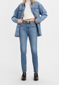 Vaqueros de mezclilla azul claro con cintura alta y corte ajustado, combinados con un top corto blanco y una camisa de mezclilla sobredimensionada. Botines negros completan el conjunto.