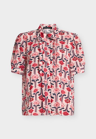 Blouse à manches courtes avec motif floral rose, rouge et marine, devant à volants et col classique sur fond clair.
