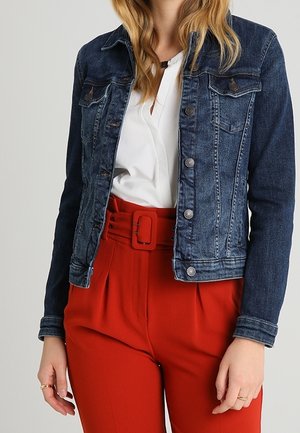 Femme portant une veste en jean bleu foncé par-dessus un chemisier blanc et un pantalon rouge taille haute avec une large ceinture.