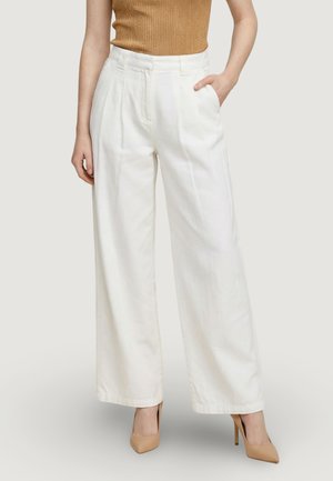 Donna che indossa pantaloni bianchi a vita alta e gamba larga con tasche laterali, abbinati a una canotta a coste senza maniche color beige e tacchi alti nudi.