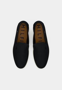 Zwarte instapschoenen met een glad leren bovenwerk, contrasterende beige zool en een bruine binnenzool waarop de tekst "HILFIGER" in marineblauw is gedrukt.