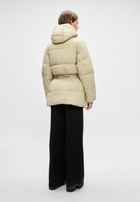 Beige puffjacka med huva, med kviltade sektioner och ett svart märkesbälte. Modellen bär svarta vida byxor och chunky stövlar.