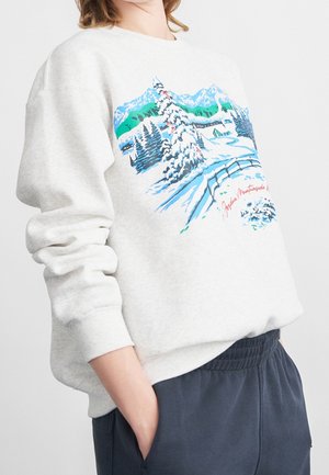 Osoba ubrana w jasnoszarą bluzę z kolorowym zimowym pejzażem górskim oraz granatowe spodnie, z rękami w kieszeniach, stojąca na białym tle.