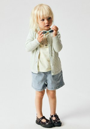 Plavokosi dječak nosi lagani džemper, žutu majicu, traper kratke hlače i crne cipele, drži sunčane naočale blizu lica, blago gleda u stranu.