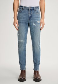 Mężczyzna ubrany w jasnoniebieskie, przetarte slim-fit dżinsy włożone do brązowych skórzanych butów, stojący na jednolitym tle.