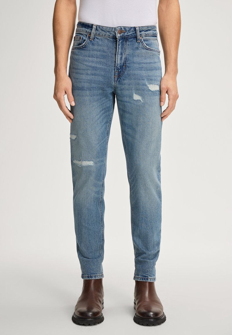 Mężczyzna ubrany w jasnoniebieskie, przetarte slim-fit dżinsy włożone do brązowych skórzanych butów, stojący na jednolitym tle.