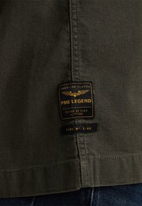 Tissu texturé vert foncé avec étiquette de vêtement PME Legend noire et jaune indiquant "American Classic, Tested at High Altitude, Type No: C-46."