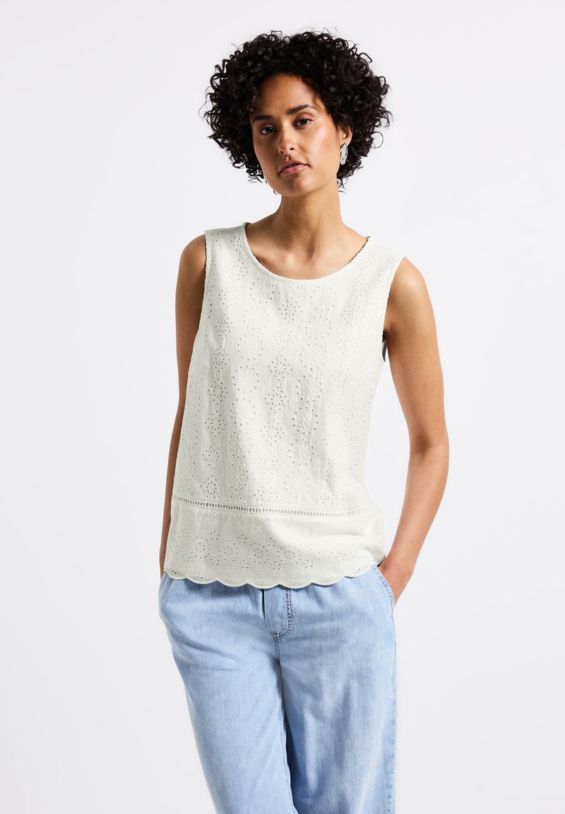Vit ärmlös blus med hålmönster, scalloped fåll och rund halsringning, kombinerad med ljusblå avslappnade jeans.