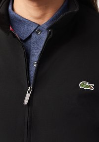 Personne portant une chemise bleue à col sous une veste noire partiellement zippée avec un petit logo crocodile vert sur la poitrine.