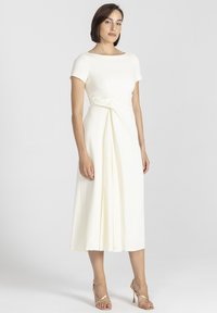 Biała midi sukienka z krótkimi rękawami, ozdobnym szczegółem w pasie, gładką teksturą tkaniny i zwiewnym dołem, połączona ze złotymi sandałami na obcasie.
