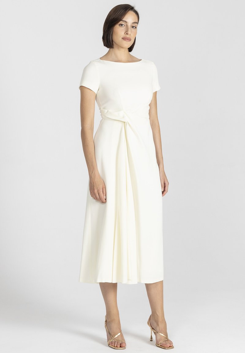 Biała midi sukienka z krótkimi rękawami, ozdobnym szczegółem w pasie, gładką teksturą tkaniny i zwiewnym dołem, połączona ze złotymi sandałami na obcasie.