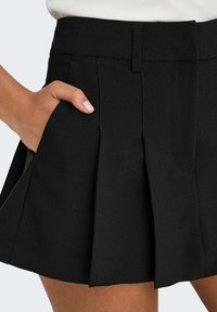Zwarte plooienrok met een tailleband, zijzakken en een gladde, gestructureerde stof. De zoom is boven de knie, wat de vorm benadrukt.