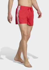 Rote Badeshorts aus leichtem Material, mit weißen Streifen an den Seiten, einem elastischen Taillenbund mit Kordelzug und einem kurzen Schnitt.