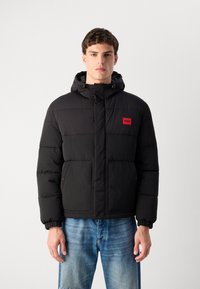 Giacca a vento nera con cappuccio, caratterizzata da imbottitura orizzontale, un patch logo rosso e chiusura con zip. Abbinata a jeans blu.