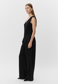 Svart linne med spetsdetaljer i kanten, tillsammans med vida svarta byxor som har en slät yta och framträdande sid sömmar, stiliserat med spetsiga svarta klackar.