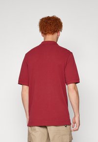 Polo majica kratkih rukava u burgundskom tonu s rebrastim ovratnikom, od pamučnog materijala, opuštenog kroja, prikazuje stražnji pogled nositelja.