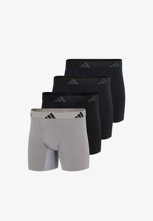 Athletischer Mann, barfuß, trägt hellgraue Adidas-Boxershorts mit schwarzem Logo am Bund vor einfarbigem Hintergrund.