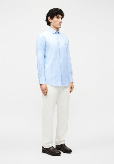 Άνδρας με σγουρά σκούρα μαλλιά, ντυμένος με ανοιχτό μπλε ριγέ πουκάμισο με κουμπιά, λευκό παντελόνι και καφέ σκούρα παπούτσια, στέκεται μπροστά σε λευκό φόντο.