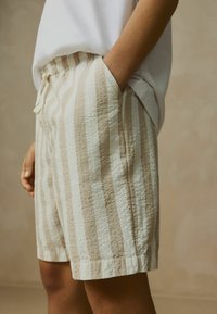 Shorts rayés beige et crème en tissu texturé, avec une taille élastique et des poches latérales. Coupe décontractée, atteignant le milieu de la cuisse.
