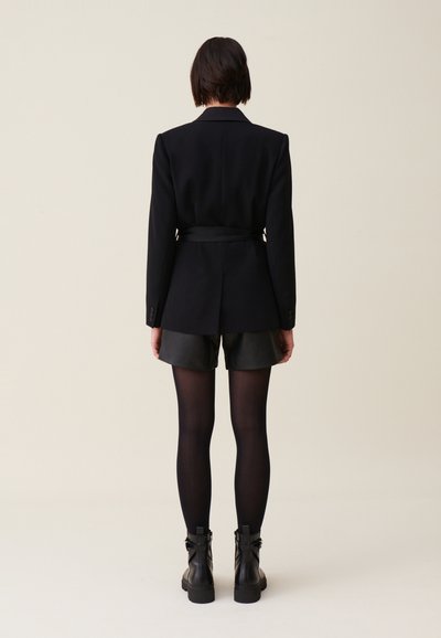Blazer strutturato nero con vita aderente, abbinato a pantaloni corti di pelle nera e collant opachi, indossato con stivaletti.