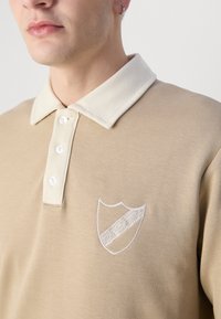 Tricou polo bej cu guler crem, având trei butoane și un logo brodat în alb cu detalii diagonale.