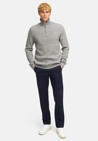 Maglione grigio a costine con colletto alto e mezza zip, abbinato a pantaloni blu scuro e sneakers bianche su uno sfondo bianco.