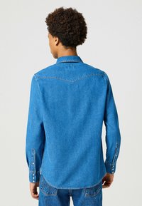 Camicia di jeans di un vivace blu, con colletto a punta, maniche lunghe con polsini a bottone e orlo curvo con dettagli di cucitura in contrasto.
