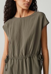 Olivegrünes, lockeres Kleid mit Rundhalsausschnitt, kurzen Kappenärmeln und Kordelzug-Akzenten in der Taille. Strukturierter Stoff.