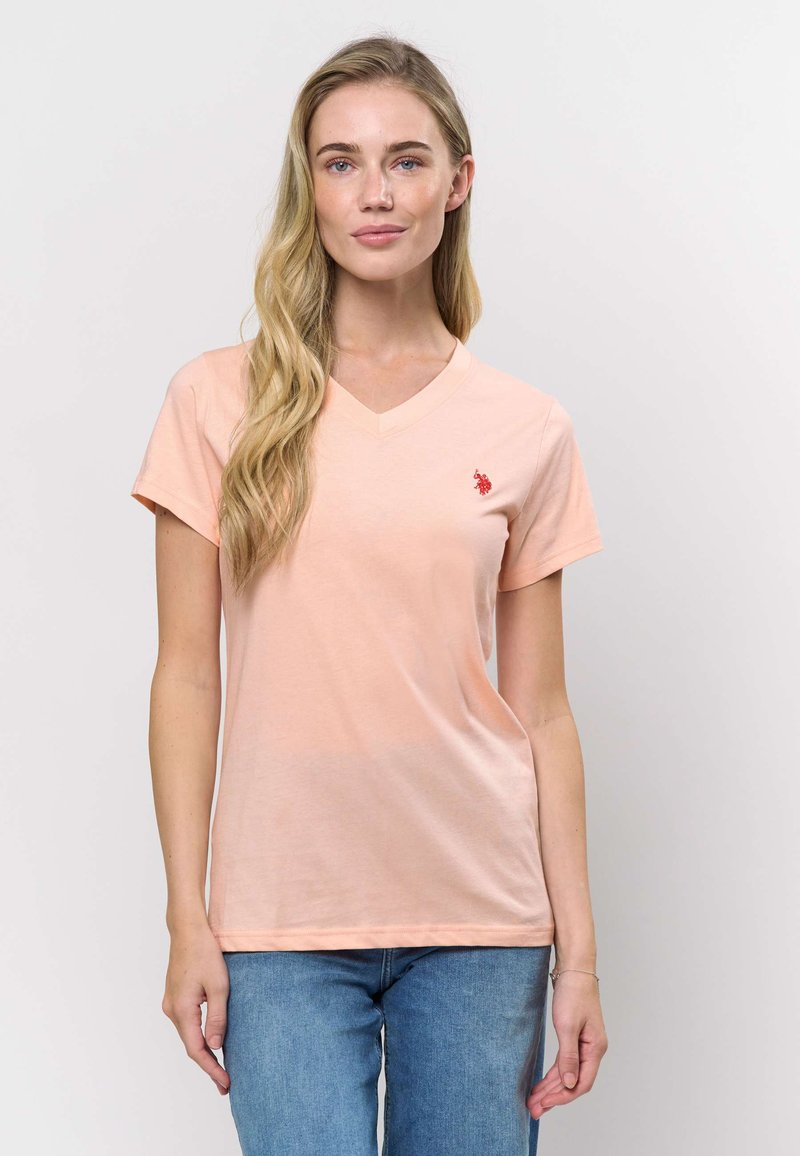 Pfirsichfarbenes V-Ausschnitt-T-Shirt aus weichem Stoff, mit kurzen Ärmeln und kleinem rotem Logo auf der linken Brust. Kombiniert mit blauen Jeans.