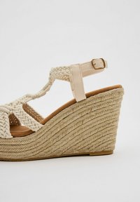 Sandalia de cuña con parte superior de cuerda trenzada en beige, plantilla marrón y una correa clara con un acento de hebilla dorada. Tacón texturizado y multicapa.
