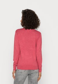 Femme aux cheveux bouclés portant un pull rose à manches longues et un jean gris, debout face à un fond blanc uni.
