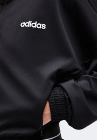 Κοντινό πλάνο μαύρου μπουφάν Adidas με λευκό λογότυπο στο στήθος και χέρι μέσα σε ελαστική τσέπη με μανσέτα.