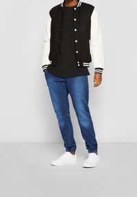 Chaqueta varsity en blanco y negro con puños rayados; camiseta negra debajo; vaqueros azules; zapatillas blancas. Diseño simple y casual.