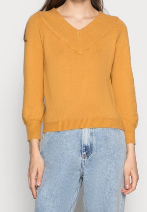 Femme portant un pull jaune moutarde à manches longues avec un col en V et un jean taille haute bleu clair sur un fond uni.