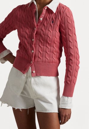 Persona che indossa un cardigan rosa a trecce sopra una camicia bianca con colletto abbinata a pantaloncini bianchi con orlo sfrangiato.