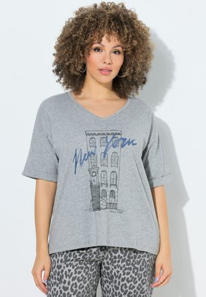Femme aux cheveux bouclés portant un t-shirt gris avec une esquisse d'un bâtiment de New York et du texte, associée à un pantalon gris à imprimé léopard, debout contre un mur blanc.