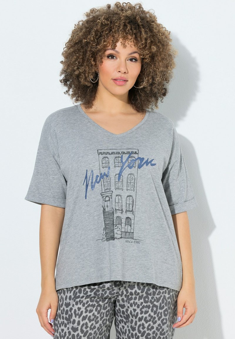 Mujer con cabello rizado que lleva una camiseta gris con un dibujo de un edificio de Nueva York y texto, combinada con pantalones grises con estampado de leopardo, de pie contra una pared blanca.