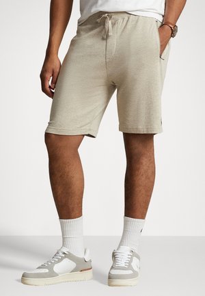 Mann trägt beige Shorts mit Kordelzug, weiße Socken sowie weiße und graue Sneakers und steht mit den Händen in den Taschen.