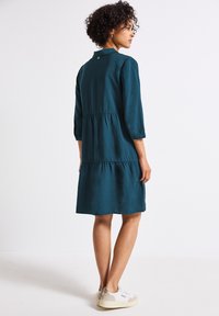 Robe teal, longueur genou, avec des manches trois-quarts, en tissu texturé, au design à volants, et avec une fermeture à bouton au dos. Portée avec des baskets décontractées.