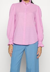 Light pink long-sleeve blouse with ruffled collar and cuffs. Textured fabric features small raised dots. Front button closure.