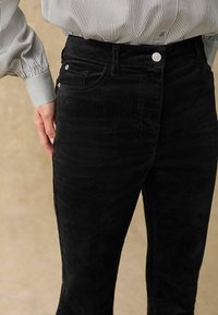 Pantalons en velours côtelé noir avec une coupe slim, dotés d'une fermeture à bouton et de poches latérales. La texture est douce avec des rayures verticales prononcées.