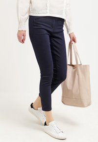 Pantalones ajustados azul marino en una tela elástica, combinados con zapatillas blancas y un bolso tote de color beige claro, con un diseño minimalista.