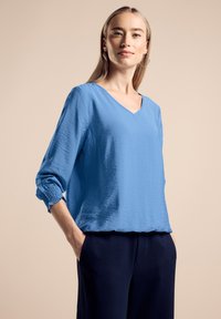 Top blu con scollo a v e maniche lunghe caratterizzato da polsini arrangiati; realizzato in un tessuto strutturato. Indossato con pantaloni blu scuro.