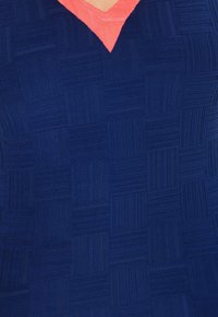 Top de tela texturada en color marino con escote en V color coral, que presenta un patrón de cuadrícula con secciones elevadas y planas para añadir dimensión.